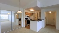 This single-floor residence offers a thoughtfully designed layout centered around an open-concept kitchen and living area. The kitchen is a functional and attractive space, featuring warm honey-toned wood cabinetry, a stainless steel gas range, a full-size refrigerator, and an under-cabinet range hood. A peninsula with a dark granite-style countertop and a built-in sink extends into the living area, creating a natural breakfast bar and defining the boundary between the kitchen and the carpeted living space. Ceiling-mounted flush lighting in the kitchen provides consistent illumination, while a pendant light with a classic glass shade adds a decorative touch over the adjacent area. The living space is finished with plush, neutral-toned carpeting that adds warmth underfoot. A mirrored wall panel in the living area reflects the kitchen and surrounding rooms, enhancing the sense of openness and amplifying natural light throughout. The layout is practical and cohesive, with clean white walls, standard ceiling heights, and smooth transitions between the kitchen's hardwood-style flooring and the carpeted common areas. Overall, the space presents a clean, move-in-ready aesthetic suited for comfortable everyday living.