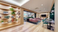 This thoughtfully designed two-floor space offers a generous and versatile layout across two well-appointed levels. The ground floor welcomes visitors with wide-plank light hardwood flooring, warm natural wood millwork, and illuminated open shelving adorned with plants and books. A central lounge area features plush pink sofas arranged around a coffee table, while an adjacent long communal workspace is lined with tufted gray banquette seating and teal accent chairs set against a deep forest green wall. A wall-mounted television is integrated into the built-in shelving, adding a functional media element. Intimate private booths, enclosed by glass sliding doors and wrapped in rich green paneling with velvet gray bench seating, provide quiet meeting or work areas. A compact kitchenette sits beneath a sweeping staircase, which ascends dramatically through a double-height atrium filled with natural light from tall windows. The upper level overlooks the ground floor through sleek black-railed balustrades and houses additional enclosed rooms with glass frontage. Throughout, a refined palette of warm wood, deep green, and soft blush creates a modern yet welcoming professional ambiance.