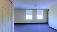 This single-floor space offers a straightforward and functional layout with a clean, understated aesthetic. The main room is generously proportioned, featuring wall-to-wall carpeting in a muted blue-gray tone that adds a soft, comfortable feel underfoot. The walls are finished in a light neutral hue, contributing to a bright and open atmosphere throughout the space. Two windows fitted with roller shades allow for adjustable natural light, keeping the interior well-lit while offering privacy as needed. The ceiling is smooth and painted white, lending an airy quality to the room, and is outfitted with a ceiling-mounted light fixture. Dark baseboard trim provides a subtle contrast against the light walls, adding a clean, defined finish at floor level. Electrical outlets are evenly distributed along the walls, offering practical utility for a variety of uses. The overall design is minimal and versatile, making the space well-suited for a range of purposes. Its open layout, neutral palette, and functional features create an environment that is easy to configure and adapt to different needs.
