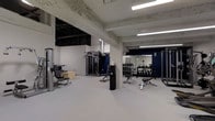 Located at 1530 Locust Street in the heart of Philadelphia, PA, this fitness center offers a well-equipped, single-floor workout facility designed to meet a range of strength and conditioning needs. The space is characterized by its industrial aesthetic, featuring exposed concrete beams and ceilings that lend a raw, urban character to the environment. Bright fluorescent strip lighting runs across the ceiling, ensuring the entire floor is evenly and thoroughly illuminated. The flooring is a clean, light grey rubberized surface well-suited for gym use. The open layout provides ample room to move freely between equipment stations. A dedicated strength training area is equipped with a cable machine and various resistance apparatuses, while a well-organized free weight section features a full dumbbell rack and adjustable benches. A large wall-mounted mirror in the free weight area enhances the sense of space and allows users to monitor their form. A bold navy blue accent wall adds a sharp visual contrast to the otherwise neutral white-and-grey color palette. The space is clean, functional, and purposefully designed to support an effective training environment.