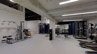 Located at 1530 Locust Street in the heart of Philadelphia, PA, this fitness center offers a well-equipped, single-floor workout facility designed to meet a range of strength and conditioning needs. The space is characterized by its industrial aesthetic, featuring exposed concrete beams and ceilings that lend a raw, urban character to the environment. Bright fluorescent strip lighting runs across the ceiling, ensuring the entire floor is evenly and thoroughly illuminated. The flooring is a clean, light grey rubberized surface well-suited for gym use. The open layout provides ample room to move freely between equipment stations. A dedicated strength training area is equipped with a cable machine and various resistance apparatuses, while a well-organized free weight section features a full dumbbell rack and adjustable benches. A large wall-mounted mirror in the free weight area enhances the sense of space and allows users to monitor their form. A bold navy blue accent wall adds a sharp visual contrast to the otherwise neutral white-and-grey color palette. The space is clean, functional, and purposefully designed to support an effective training environment.