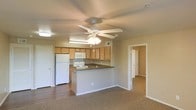 This single-floor space is designed with an open-concept layout that seamlessly connects the kitchen and living area. The kitchen is equipped with light wood cabinetry, a white refrigerator, a white range, and a built-in microwave. A peninsula counter with a dark laminate surface extends into the living area, providing additional workspace and serving as a natural divider between the two zones. The living area features carpeted flooring, while the kitchen has hard surface flooring, creating a practical distinction between the spaces. A ceiling fan with a built-in light fixture is centrally positioned overhead, providing both airflow and ambient lighting throughout the open area. The neutral wall tones and standard ceiling height contribute to a clean, functional aesthetic. Additional rooms are accessible through doorways visible from the main living space, suggesting further living or storage areas beyond the open floor plan. The overall design prioritizes practicality and ease of movement, making efficient use of the available space throughout the unit.