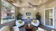 This single-floor community clubhouse is thoughtfully designed to blend comfort and functionality in an inviting, resort-style setting. The main lounge area serves as the centerpiece of the space, featuring elevated ceilings with a modern ceiling fan, recessed lighting, and a wall-mounted flat-screen TV. White upholstered armchairs and sofas are arranged around a circular wood-and-glass coffee table, accented by colorful throw pillows in green, yellow, and blue tones. A decorative stone-faced media console anchors the seating area, flanked by tall tropical potted plants that add a lush, organic element to the interior. Large floor-to-ceiling windows wrap around the lounge, flooding the space with natural light and framing views of the well-maintained courtyard and surrounding landscaping. The game room offers a full-sized pool table as its focal point, set against vibrant green walls and overlooking the community pool area through a large picture window — a bright and energetic contrast to the lounge's relaxed atmosphere. Glass-paneled entry doors connect the interior to an outdoor patio area, reinforcing the seamless indoor-outdoor flow. The combination of neutral wall tones, patterned carpeting, and natural greenery gives the entire space a warm, welcoming ambiance ideal for socializing and leisure.
