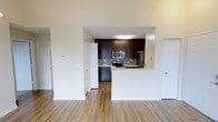 This single-floor residence offers a clean, modern design with an open and airy feel. The space centers around an open-concept kitchen and living area, where light-toned hardwood-style flooring runs seamlessly throughout, adding warmth and visual continuity.

The kitchen is a standout feature, showcasing rich dark espresso cabinetry that extends to the upper walls, complemented by a neutral mosaic tile backsplash that adds texture and depth. Stainless steel appliances — including a refrigerator, range, and microwave — provide a sleek, contemporary finish. A white peninsula counter extends outward, creating a natural division between the kitchen and the living area while offering additional workspace and casual seating potential.

The layout is thoughtfully designed with multiple doorways visible from the main living area, suggesting well-distributed access to additional rooms and storage. The tall, smooth white walls and flush ceiling lighting contribute to a bright, clean atmosphere throughout.

Overall, the space delivers a functional yet stylish environment, balancing modern kitchen aesthetics with open living areas. The neutral color palette of whites, warm wood tones, and dark cabinetry creates a timeless and inviting ambiance well-suited for everyday living.