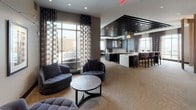 Located at 4800 Auburn Ave in Bethesda, Maryland, this sophisticated amenity lounge is a well-appointed communal space designed for both relaxation and socializing. The single-floor layout is thoughtfully organized into distinct functional zones, each with its own character while maintaining a cohesive contemporary aesthetic throughout.

The lounge opens into a refined seating area furnished with tufted swivel chairs and a round coffee table, accented by framed artwork on neutral walls and floor-to-ceiling windows dressed in bold patterned drapes. These large windows flood the space with natural light and provide views of an outdoor terrace.

A separate lounge zone features rich, dark leather sofas and armchairs arranged around coffee tables, illuminated by striking sculptural gold pendant lights. Floor-to-ceiling glass doors open directly to the terrace, reinforcing the connection between indoor and outdoor spaces.

At the heart of the space, a sleek bar with a white countertop and dark cabinetry serves as an entertaining hub, complemented by bar-height seating. Dramatic linear ceiling details with bold dark beams and elegant pendant fixtures define this area.

A dedicated game room offers a billiards table, wall-mounted television, and bar stools, creating an inviting recreational retreat. The entry corridor features a classic checkered tile floor and dark-paneled walls, providing elevator and area-of-refuge access, completing this upscale amenity floor.