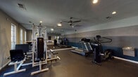 This fitness center offers a dedicated space for exercise and wellness within a single-floor layout. The room features durable rubber flooring throughout, providing excellent traction and impact absorption for a variety of workouts. A striking multi-toned accent wall — transitioning from deep navy at the base through graduated shades of gray — gives the space a modern, polished aesthetic.

The gym is equipped with a multi-station cable weight machine, ideal for a comprehensive strength training routine, along with a weight bench and additional free weight accessories. For cardio, the space includes multiple treadmills and an elliptical cross-trainer, offering users versatile options for aerobic exercise. A wall-mounted water fountain is conveniently available within the room.

A large wall mirror extends along one side of the space, visually expanding the room and allowing users to monitor their form during workouts. Recessed lighting and a ceiling fan with an integrated light fixture ensure the space remains well-lit and well-ventilated. Natural light filters in through windows near the entrance, adding brightness to the overall atmosphere. Access is controlled via a key-fob entry door, and posted fitness center rules are displayed near the entrance. The space is compact yet thoughtfully arranged to maximize utility and comfort for users.