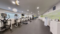 Located at 2100 Flintshire Rd in Rosedale, MD, this single-floor fitness center is designed to provide a well-rounded workout environment. The space is laid out in a long, open floor plan with dark rubber flooring throughout, offering durability and comfort underfoot during exercise.

The cardio section is equipped with multiple treadmills and elliptical machines, positioned along one side of the room for easy access. Natural light filters in through window blinds, complementing the recessed ceiling lighting that illuminates the entire facility. A wall-mounted flat-screen television is positioned near the cardio equipment for entertainment during workouts.

Toward the rear of the space, a strength training area houses a cable/multi-station machine, a full dumbbell rack with a weight bench, and various additional fitness accessories including stability balls. A large wall mirror in this section enhances the sense of space and allows users to monitor their form.

Motivational words and phrases — such as "limitless," "determination," "inspire," and "perseverance" — are displayed prominently in green and white wall graphics, contributing to an energizing atmosphere. A water cooler and a check-in station with white cabinetry are conveniently placed along one wall. The facility also includes a restroom with a wood-toned vanity, white countertop, and framed mirror.