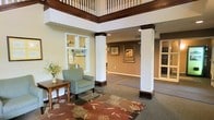 This inviting lobby space makes a strong first impression with its thoughtful blend of classic architectural design and warm, comfortable furnishings. Prominent white columns with rich dark wood bases and crown details anchor the space, drawing the eye upward to an elegant upper-level balcony framed by traditional wood balusters and a bold dark wood rail — a standout architectural feature that lends the space a sense of height and openness. The flooring transitions between carpet and a decorative area rug with floral motifs in warm earth tones, adding visual interest and a welcoming feel underfoot. Comfortable seating, including a sage green armchair and a classic wooden side table adorned with a fresh floral arrangement, create a relaxed reception area. Framed botanical and nature-inspired artwork lines the walls, contributing to the space's refined yet approachable character. Recessed lighting and wall sconces provide a soft, well-distributed glow throughout. Beyond the main seating area, the space opens into additional corridors and rooms, suggesting a larger facility designed for comfort and easy navigation. Overall, the lobby exudes a warm, professionally appointed atmosphere well-suited to welcoming visitors and residents alike.