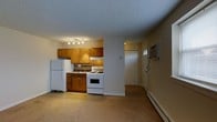 This single-floor space offers a functional and straightforward layout that combines the kitchen and living area in an open-concept arrangement. The kitchen is equipped with warm honey-toned wood cabinetry, a white gas range with oven, a full-size refrigerator, and a sink set beneath a tile backsplash. A track lighting fixture overhead provides direct illumination to the cooking and prep areas. The textured popcorn ceiling adds a classic character throughout the space. Adjacent to the kitchen, the living area offers an open and versatile footprint, with neutral-toned flooring that extends throughout. A baseboard heater runs along the wall, providing efficient in-unit heating, while a wall-mounted air conditioning unit near the entryway ensures year-round climate comfort. The entryway features a solid paneled door with a sidelight window, allowing a touch of natural light into the foyer area. The overall ambiance is practical and understated, offering a clean canvas well-suited for personalization. The space is designed with everyday livability in mind, balancing essential amenities within a compact and efficient footprint.
