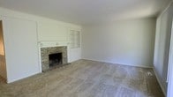 This two-story home presents a traditional and functional layout designed to offer comfortable living across multiple levels. The main living area is a notable highlight, centered around a classic brick fireplace with a white-painted wood mantel that serves as the room's focal point. Flanking the fireplace is built-in cabinetry with glass-paneled doors, providing both display and storage potential while reinforcing the space's traditional character. The room is generously proportioned, offering ample space for a variety of furniture arrangements. Neutral-toned wall-to-wall carpeting runs throughout the living area, contributing to a warm and understated atmosphere. The walls are finished in a crisp, light neutral tone that reflects available light and lends a clean, airy feel to the interior. The overall design aesthetic is classic and straightforward, with architectural details that suggest a well-established build. The two-floor layout provides the opportunity for clear separation between living and private spaces, adding to the home's practicality and livability.