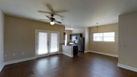 Located in Denton, Texas, this single-story space offers a well-designed, open-concept layout that seamlessly connects the living area and kitchen. Rich, dark hardwood flooring runs throughout, providing a warm and polished foundation that contrasts beautifully with the neutral beige walls and crisp white trim.

The living area is bright and airy, benefiting from multiple windows dressed with white horizontal blinds that allow for generous natural light. A ceiling fan with a built-in light fixture adds both comfort and functionality to the space. A set of French doors provides an additional source of natural light and offers access to the exterior.

The kitchen is thoughtfully integrated into the open floor plan and features dark cabinetry, stainless steel appliances including a refrigerator and microwave, and a peninsula-style counter that serves as a natural divider between the kitchen and living areas. A pendant light hangs above the dining area, adding a refined touch.

Overall, the space presents a clean, modern aesthetic with a cohesive color palette, practical finishes, and a layout that maximizes flow and functionality. The combination of natural light, quality flooring, and contemporary kitchen design makes this a comfortable and inviting environment.