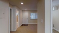 This single-floor space presents a clean, functional layout with a calm and neutral aesthetic throughout. The entryway serves as the focal point of the main area, featuring a classic white paneled door accented by a small entry mat, setting a welcoming tone upon arrival. Recessed ceiling lights provide warm, even illumination across the space, complementing the soft beige carpeted flooring that extends throughout the room. The walls are finished in a warm neutral tone, contributing to a cohesive and understated atmosphere. A window fitted with white horizontal blinds allows for natural light to filter in while maintaining privacy, brightening the space without overwhelming it. White baseboards and trim offer crisp contrast against the warm wall color, adding a polished finish to the interior. An open doorway to the side leads to an adjoining room — visible from the main area — which appears to be a bathroom, featuring lighter tile or flooring. The overall layout is straightforward and practical, with clear sightlines and easy flow between areas. The space is ideal for those seeking a no-frills, move-in ready environment with a neutral palette that accommodates a wide range of furnishing and décor styles.
