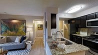 This single-floor residence offers a thoughtfully designed open-concept layout that connects the kitchen, living area, and hallway in a cohesive and functional arrangement. The kitchen serves as a central highlight, featuring granite countertops with an integrated stainless steel sink, dark cabinetry, stainless steel appliances including a full-size refrigerator and microwave, and a peninsula that doubles as a breakfast bar — ideal for casual dining or meal prep. The flooring throughout the main living areas consists of light-toned wood-style planks that lend warmth and visual continuity to the space. The living area is comfortably scaled, offering room for a full seating arrangement, and is accented by a textured ceiling that adds subtle architectural character. A well-lit hallway connects the main living spaces to the additional rooms, creating a smooth flow throughout the home. Warm, ambient lighting from a combination of flush-mount ceiling fixtures and floor lamps contributes to an inviting atmosphere. The neutral color palette of the walls and furnishings provides a versatile backdrop that is easy to personalize, making this a practical and welcoming space suited for everyday living.
