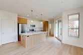 This thoughtfully designed three-story home offers 3 bedrooms, 4 bathrooms, and a spacious attached garage across three distinct levels. The ground floor houses the garage, providing convenient direct-access parking. The main living level features a generous open-concept kitchen and living area anchored by a large center island with a white quartz countertop, a dual basin sink, and pendant lighting overhead. Cabinetry combines white upper cabinets with warm wood-toned lower cabinets and a full suite of stainless steel appliances, including a French door refrigerator, electric range, and built-in microwave. Light hardwood flooring runs throughout this level, and a sliding glass door opens to an outdoor area, allowing natural light to fill the space. The upper floor is dedicated to the bedrooms, including a spacious primary suite with plush carpeting, a walk-in closet with wire shelving, and an en-suite bathroom. The bathrooms throughout feature clean, modern finishes including wood-grain vanities, white quartz countertops, large mirrors, and tile flooring. A staircase with a warm wood handrail connects all three levels, complemented by bright, white walls and an airy, open feel throughout the home.