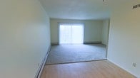 This single-floor space features an open living area that combines two distinct flooring types: smooth hardwood-style flooring near the entry transitions seamlessly into plush carpeting that covers the main portion of the room. The layout feels open and unobstructed, offering a sense of spaciousness well-suited for flexible furniture arrangements.

A large sliding glass door dominates the far wall, fitted with vertical blinds that diffuse natural light softly throughout the room, creating a bright and airy atmosphere. The door likely provides access to an outdoor area, adding both functionality and visual openness to the space.

The walls are finished in a neutral, light tone that complements the natural light and contributes to the room's clean, understated aesthetic. Baseboard heating units run along multiple walls, ensuring efficient and even heat distribution throughout. The ceiling is smooth and neutral, maintaining a simple, uncluttered look overhead.

Overall, the space presents a functional and well-lit interior with practical features, neutral finishes, and a straightforward layout that offers versatility for a variety of uses.
