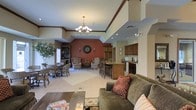 This single-floor space features an open-concept layout that seamlessly connects multiple functional zones, creating a welcoming and versatile environment. The interior is characterized by vaulted ceilings with decorative crown molding and angled architectural details that add visual interest and a sense of height throughout the space.

The living area is anchored by a plush, earth-toned sofa accented with decorative throw pillows, positioned around a rustic wooden coffee table atop a patterned area rug. A chandelier with warm lighting serves as a central focal point overhead.

Adjacent to the living area, a casual seating arrangement with upholstered armchairs and a decorative accent table rests on a second area rug, providing an intimate conversation space. A bold terracotta-red accent wall with wall sconces and a circular decorative piece adds warmth and visual contrast.

The dining area features a wooden table with bar-height seating, transitioning naturally into a kitchen space outfitted with dark wood cabinetry, a countertop peninsula, and standard appliances including a microwave. A separate dining nook near large windows offers additional seating with natural light. A built-in bookcase along one wall adds functional storage and character to the overall space.