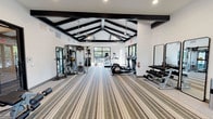Located at 3501 W Walnut Hill Ln in Irving, TX, this well-appointed fitness center offers a thoughtfully designed single-floor layout ideal for a full range of workout routines. The space is immediately distinguished by its dramatic vaulted ceiling adorned with striking black exposed beams, which lend a modern yet structural character to the interior. Ceiling fans are suspended from the peaks, ensuring comfortable airflow throughout the facility.

The flooring throughout is a striped, multi-tone carpet that adds visual texture while providing underfoot comfort during training sessions. Large black-framed windows and glass doors line multiple walls, flooding the space with natural light and offering views of the surrounding outdoor greenery, creating an open and energizing atmosphere.

The fitness center is comprehensively equipped for both strength and cardiovascular training. Strength equipment includes a Smith machine with a weight bench, a functional trainer, cable machines, a dumbbell rack with a full range of free weights, and additional resistance machines. The cardio section features ellipticals and treadmills positioned near the glass doors with outdoor views. Full-length mirrors are strategically placed to support proper form. A granite-topped counter provides a convenient surface for personal items. The space also includes safety features such as a fire extinguisher and emergency exit signage.