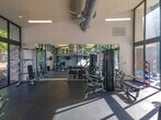 Located at 5155 S Old Peachtree Rd in Peachtree Corners, Georgia, this single-floor fitness center is designed to deliver a bright, open, and well-equipped workout environment. The space is defined by its dramatic floor-to-ceiling glass walls framed in sleek black metal, which flood the interior with abundant natural light and offer expansive views of the surrounding greenery and pool area. Soaring ceilings with exposed industrial-style ductwork add an urban, modern character to the space. The rubber-floored workout area is divided into distinct functional zones. The cardio section features a row of treadmills and elliptical machines positioned to take full advantage of the panoramic outdoor views, along with wall-mounted televisions for entertainment during workouts. The strength training area is equipped with a multi-station cable machine, adjustable benches, a full rack of dumbbells, and a dedicated free weights zone, all complemented by large wall-to-wall mirrors that visually expand the space and support proper form. Natural light streams across the dark rubber flooring throughout the day, creating an energizing atmosphere. The facility offers direct views of an outdoor pool, further enhancing the overall experience of this well-appointed community fitness center.