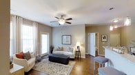 This single-floor residence offers a thoughtfully designed layout that balances comfort and functionality. The living area serves as the heart of the space, showcasing rich hardwood flooring that extends throughout and warm, neutral wall tones that create a welcoming ambiance. A ceiling fan with a light fixture anchors the room, providing both airflow and soft illumination to complement the natural light streaming in through multiple windows dressed with elegant white curtains. The living area offers generous room for seating and relaxation, with an open sightline that connects effortlessly to adjacent spaces. A doorway leads to what appears to be a well-lit bathroom, hinting at a practical and accessible floor plan. The open-concept flow continues toward a dining or bar area, visible at the edge of the room, adding versatility to the layout. Neutral-painted walls paired with white trim and door frames give the space a clean, polished look throughout. The overall ambiance is bright, airy, and inviting — a residence that combines classic design elements with everyday livability in a cohesive and comfortable single-level layout.