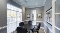 This single-floor space functions as a well-appointed meeting or conference room, thoughtfully designed for focused gatherings and professional discussions. The room is finished in a cool, medium-toned grey on the walls, complemented by crisp white crown molding and a chair-rail detail that adds a refined architectural touch. A round dark-finished table serves as the centerpiece, surrounded by four black executive-style chairs that provide both comfort and a polished aesthetic.

Natural light is a defining feature of the space, with multiple windows fitted with white horizontal blinds that allow for flexible light control while maintaining a clean, consistent look. The windows offer views of lush greenery outside, contributing to a calm and inviting atmosphere. Recessed lighting and a ceiling fan are integrated into the tray ceiling above, ensuring the room remains well-lit and comfortable regardless of the time of day. Framed artwork is displayed on the walls, adding a curated visual element without overwhelming the space. Neutral carpeting grounds the room and completes its professional, welcoming character.