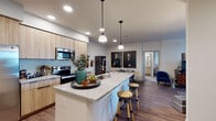 This single-floor residence showcases a thoughtfully designed open-concept layout with modern finishes and a cohesive contemporary aesthetic. The kitchen serves as a central highlight, featuring light wood cabinetry that extends to the ceiling, stainless steel appliances including a French door refrigerator, electric range, and built-in microwave. A decorative blue geometric tile backsplash adds a distinctive visual element, while the expansive granite-topped island provides generous counter space, an undermount sink, and seating for multiple guests via wooden bar stools. Pendant lighting over the island complements the recessed ceiling lights throughout. Wood-look flooring flows seamlessly from the kitchen into the open living area, which offers sliding glass door access to a private balcony, filling the space with natural light. The bedrooms are carpeted for comfort, with windows dressed in patterned curtains that provide both style and privacy. Both bathrooms echo the kitchen's design language, featuring the same light wood vanity cabinetry paired with granite countertops and large mirrors. One bathroom includes a walk-in glass-enclosed shower, while the second offers a shower-tub combination. The overall ambiance is modern and refined, with a neutral color palette accented by curated décor throughout.