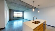 Located at 122 Delaware St in Kansas City's historic River Market district, this modern urban unit showcases an industrial-chic aesthetic defined by polished concrete floors and dramatic exposed concrete ceilings throughout. The open-concept living and kitchen area is anchored by a sleek island with a white quartz countertop, double stainless steel sink, warm wood cabinetry, and pendant lighting. The kitchen is fully equipped with stainless steel appliances, white upper cabinets extending to the ceiling, and in-unit stacked washer and dryer hookups conveniently tucked nearby. Large black-framed windows flood the living spaces with abundant natural light while framing impressive views of the Missouri River and surrounding greenery. The bedroom continues the industrial theme with exposed concrete ceilings and equally generous window views of the river landscape. The bathroom features polished concrete floors, clean white tile surrounding a full soaking tub with shower, and a minimalist design consistent with the rest of the unit. The thoughtful combination of raw industrial materials and contemporary finishes creates a distinctive, loft-inspired atmosphere well-suited for urban living in Kansas City.