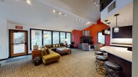 Located at 10555 SE Mather Rd in Clackamas, Oregon, this two-story community amenity building delivers a dynamic and thoughtfully designed environment with a bold, contemporary aesthetic. The main level opens into a spacious lounge area characterized by soaring vaulted ceilings, track lighting, and an expansive wall of windows that floods the space with natural light and frames lush outdoor greenery. A striking orange accent wall anchors the lounge, which features a tile-surround fireplace, plush sectional seating, and colorful accent chairs arranged for conversation and relaxation. An adjacent dining area with high-top tables and yellow leather chairs adds a lively social hub, complemented by a decorative wood-panel accent wall and hardwood flooring. A well-appointed kitchen offers dark espresso cabinetry, granite-style countertops, stainless steel appliances, and warm wood-look flooring, ideal for community gatherings. A dedicated media room showcases vaulted ceilings with exposed wood beam detailing, skylights, an orange sectional sofa, a large flat-screen display, and framed vintage movie posters for a fun cinematic atmosphere. The lower level houses a fitness center equipped with treadmills and elliptical machines, rubber flooring, and garden-facing windows that bring in natural light. A second-floor mezzanine with a wrought-iron railing overlooks the entry level, adding architectural depth throughout.