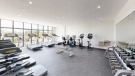 Located at 1 Curtiss Pky in Miami Springs, Florida, this fitness center offers a dedicated exercise room designed to support a comprehensive workout routine. The space is finished with a clean, modern aesthetic — bright white walls, recessed lighting, and dark rubberized flooring that provides both durability and comfort underfoot.

Large floor-to-ceiling windows line one wall, flooding the room with natural light while offering open views of the surrounding outdoor environment. The generous amount of natural light enhances the airy, motivating atmosphere of the space.

The exercise room is well-appointed with a wide array of equipment. Cardio options include multiple treadmills, stationary bikes, elliptical trainers, and stair climbers, catering to a variety of fitness preferences. A full dumbbell rack with an extensive range of free weights is positioned alongside a flat workout bench, supporting strength training needs. A large wall mirror reflects both the equipment and the natural light, adding to the sense of openness.

The layout is thoughtfully organized to allow ample space between machines, ensuring ease of movement throughout the room. A water station is also available for convenience. The overall ambiance is clean, functional, and inviting, making this fitness center a practical and well-rounded space for everyday exercise.