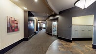 This single-floor lobby presents a polished and contemporary design that makes a strong first impression. The entrance foyer is bright and welcoming, drawing in natural light through large floor-to-ceiling windows that overlook the exterior streetscape. A seating area near the entry features modern upholstered furniture, including a sofa and armchairs arranged around a bold red accent table, providing a comfortable space for residents and guests.

The architectural design is defined by sweeping curved walls and tiered ceiling details finished in contrasting warm gold and deep brown tones, lending the space a dynamic, upscale character. Recessed lighting throughout maintains a warm, ambient glow that complements the dark wood wall panels and charcoal-toned base tiles.

The main corridor extends from the lobby with patterned carpet flooring and continues past a dedicated fitness center, visible through a glass-paneled door and equipped with exercise machines. Wall-mounted sconces provide accent lighting along the hallway, and decorative artwork adds visual interest to the passage.

A dedicated mailroom alcove houses a full bank of individual resident mailboxes. Adjacent to the lobby, a pair of stainless steel elevators with dark tile surrounds offer efficient vertical access. A prominent pendant light fixture anchors the elevator landing area, completing the cohesive and refined aesthetic of this well-appointed building lobby.