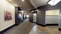 This single-floor lobby presents a polished and contemporary design that makes a strong first impression. The entrance foyer is bright and welcoming, drawing in natural light through large floor-to-ceiling windows that overlook the exterior streetscape. A seating area near the entry features modern upholstered furniture, including a sofa and armchairs arranged around a bold red accent table, providing a comfortable space for residents and guests.

The architectural design is defined by sweeping curved walls and tiered ceiling details finished in contrasting warm gold and deep brown tones, lending the space a dynamic, upscale character. Recessed lighting throughout maintains a warm, ambient glow that complements the dark wood wall panels and charcoal-toned base tiles.

The main corridor extends from the lobby with patterned carpet flooring and continues past a dedicated fitness center, visible through a glass-paneled door and equipped with exercise machines. Wall-mounted sconces provide accent lighting along the hallway, and decorative artwork adds visual interest to the passage.

A dedicated mailroom alcove houses a full bank of individual resident mailboxes. Adjacent to the lobby, a pair of stainless steel elevators with dark tile surrounds offer efficient vertical access. A prominent pendant light fixture anchors the elevator landing area, completing the cohesive and refined aesthetic of this well-appointed building lobby.