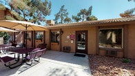 Located at 2625 E Tropicana Ave in Las Vegas, Nevada, this single-story community clubhouse serves as a central hub for residents. The building features a Southwestern-inspired stucco exterior with a clay tile roof, framed by mature pine and palm trees that provide natural shade throughout the property.

The heart of the clubhouse is a generously sized common room designed for both socializing and relaxation. The space is furnished with patterned upholstered armchairs and sofas arranged around a central coffee table, complemented by a dedicated dining area with wooden tables and chairs. Multiple ceiling fans keep the room comfortable, while drape-framed windows allow natural light to fill the interior. A flat-screen television anchors one wall, making this an ideal gathering space. Tile flooring runs throughout, accented by area rugs that define the seating zones.

An on-site laundry room provides added convenience, equipped with coin-operated washers and dryers alongside a bright window overlooking the outdoors. The exterior features a covered patio with seating and umbrella tables, ideal for outdoor relaxation. The crown jewel of the property is a sparkling resort-style swimming pool, enclosed by decorative iron fencing and surrounded by a spacious deck, with tall palms and pines creating a lush, shaded atmosphere.