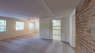 This single-floor residence offers a practical and comfortable layout designed for everyday living. The spacious main living area is finished with plush carpeting and features a ceiling fan with light fixture, creating a bright and airy atmosphere. Natural light flows in through multiple windows fitted with blinds, lending the space a warm, inviting feel. Louvered bi-fold doors open to reveal the kitchen, which is equipped with white cabinetry accented by gold-tone hardware, a gas range, ample counter space with a sink, and a refrigerator. The tile flooring in the kitchen provides a clean contrast to the carpeted living spaces. A distinctive exposed brick accent wall adds a touch of character to one section of the home. The bedrooms are generously sized, each outfitted with ceiling fans and carpeted floors. One bedroom features a row of windows that invite abundant natural light and outdoor views, while another includes a built-in closet with louvered doors for organized storage. The bathroom is clean and functional, offering a tiled tub and shower combination, a vanity with sink, and mosaic tile flooring. Throughout the space, neutral tones and consistent finishes create a cohesive and understated aesthetic.