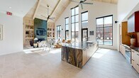 This contemporary clubhouse in Richmond, VA is designed to impress, offering a thoughtfully curated suite of amenity spaces across a single, generously sized floor. The centerpiece is a soaring open-concept great room with dramatic vaulted ceilings clad in natural wood, oversized black-framed floor-to-ceiling windows, and large-format light tile flooring. Abundant natural light pours in from multiple directions, offering views of the surrounding community. Industrial-scale ceiling fans complement the architectural height, while a striking fireplace with emerald mosaic tile and built-in shelving anchors one end of the room.

The gourmet kitchen features light wood cabinetry, a stunning waterfall-edge island with richly veined stone countertops, a professional range, and a warm salmon-toned zellige tile backsplash that adds texture and character. A separate bar counter with matching cabinetry provides additional prep and serving space.

The fitness center is well-appointed with high vaulted ceilings, exposed beams, wall-to-wall mirrors, cardio machines, strength training equipment, and ample natural light from large windows.

A private meeting room enclosed by sleek glass sliding doors features deep green accent walls, a round white conference table, tan upholstered chairs, and a pendant light fixture. Connecting hallways with glass-walled rooms and curated artwork tie the entire space together cohesively.