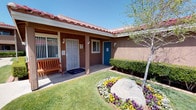 This single-story property showcases a classic Southwestern architectural style, characterized by a distinctive red clay tile roof and warm brown stucco exterior walls. The covered front entryway provides a sheltered, inviting entrance, complete with a wooden bench and wall-mounted lantern-style lighting — a thoughtful detail that adds a welcoming charm. A "Welcome Home" doormat greets visitors at the security screen door, reinforcing the warm, residential atmosphere.

The outdoor grounds are meticulously maintained, featuring lush green lawns that frame the building's exterior. A standout landscaping feature is the decorative boulder centerpiece surrounded by vibrant flower beds of yellow, white, and purple blooms, adding a striking splash of color to the natural surroundings. Neatly trimmed hedges line the building's facade, contributing to a tidy and polished appearance.

A gently curving concrete walkway winds alongside the building, bordered by well-kept grass and mature trees that provide natural shade. The overall exterior ambiance is bright and cheerful, benefiting from abundant natural sunlight under clear blue skies, creating a peaceful and welcoming environment throughout the property's outdoor spaces.