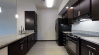 This single-floor space showcases a contemporary design with clean lines and a neutral color palette throughout. The open-concept layout connects the kitchen and living area, creating a cohesive and functional flow. Light hardwood-style flooring extends across the space, complementing the crisp white walls and dark baseboards.

The kitchen is a standout feature, outfitted with dark espresso cabinetry that provides ample storage, granite countertops, and a suite of black appliances including a refrigerator, range, and built-in microwave. A double sink with a modern faucet anchors one side of the kitchen, while a peninsula with pendant lighting defines the boundary between the kitchen and the main living area. Two sleek hanging pendant lights add warmth and visual interest overhead.

The living area benefits from natural light through a front-facing window and is equipped with a ceiling fan. The open sightlines from the entryway through to the living space give the unit an airy, expansive feel. Overall, the space balances modern aesthetics with practical functionality.