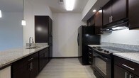 This single-floor space showcases a contemporary design with clean lines and a neutral color palette throughout. The open-concept layout connects the kitchen and living area, creating a cohesive and functional flow. Light hardwood-style flooring extends across the space, complementing the crisp white walls and dark baseboards.

The kitchen is a standout feature, outfitted with dark espresso cabinetry that provides ample storage, granite countertops, and a suite of black appliances including a refrigerator, range, and built-in microwave. A double sink with a modern faucet anchors one side of the kitchen, while a peninsula with pendant lighting defines the boundary between the kitchen and the main living area. Two sleek hanging pendant lights add warmth and visual interest overhead.

The living area benefits from natural light through a front-facing window and is equipped with a ceiling fan. The open sightlines from the entryway through to the living space give the unit an airy, expansive feel. Overall, the space balances modern aesthetics with practical functionality.