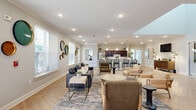 This single-floor community amenity space is thoughtfully designed to offer residents a variety of social and recreational areas within a cohesive, modern aesthetic. The open-concept main lounge serves as the heart of the space, featuring herringbone-patterned hardwood floors, recessed lighting, and abundant natural light from multiple windows with white blinds. The lounge is furnished with a mix of seating options, including a dark leather sofa, cushioned armchairs, and a round coffee table, all anchored by a patterned area rug. Decorative circular wall art adds a curated, artistic touch throughout. Adjacent to the lounge, a full kitchen offers dark wood cabinetry, stainless steel appliances, subway tile backsplash, and a long white quartz island with bar seating for several guests, illuminated by elegant pendant lights. A separate media room provides a more intimate setting with reclining lounge chairs, woven ottomans, a wall-mounted flat-screen TV, and bold abstract artwork. A small dining nook features a round wood table with cane-back chairs, ideal for casual meetings. The dedicated fitness center is equipped with a multi-function cable machine, treadmill, elliptical, stationary bike, and weight bench, all set on durable flooring with natural light from surrounding windows.