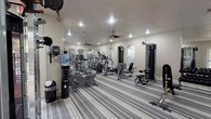 This fitness center offers a thorough range of exercise equipment spread across a single-floor layout designed to accommodate a variety of workout routines. The space is defined by its series of arched windows that line the perimeter walls, allowing natural light to flow in and providing outdoor views of surrounding greenery and common areas. Recessed lighting and ceiling fans are distributed throughout, ensuring the space remains well-lit and comfortable during use.

The cardio section is well-stocked with multiple treadmills, elliptical trainers, and stationary bikes, including Life Fitness branded equipment. A dedicated tuck of treadmills is positioned near a corner alcove of arched windows, offering a pleasant outdoor view during workouts. The strength training area includes cable pulley machines, a functional trainer, a leg press machine, and a rack of dumbbells arranged along a mirrored wall, allowing users to monitor form. A wall-mounted television is also present for entertainment.

The flooring throughout consists of striped, grey-toned carpet, lending a clean and cohesive aesthetic. A water cooler station is conveniently positioned near the entrance. French doors on one side provide direct access to an outdoor area, enhancing ventilation and connectivity to the broader facility.