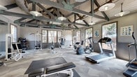 Located at 7009 Almeda Rd in Houston, TX, this dedicated fitness center offers a thoughtfully designed exercise space that combines industrial-chic aesthetics with functional workout amenities. The room's most striking architectural feature is its soaring vaulted ceiling, characterized by exposed wood planking and a network of structural beams and trusses accented in gray and green tones. Pendant lighting fixtures hang from the ceiling framework, casting warm, even illumination throughout the space. Large windows line multiple walls, flooding the interior with natural light and offering views of the surrounding outdoor greenery. The floors are covered in a geometric-patterned carpet in varying shades of gray, adding a modern visual element underfoot. Full-length mirrors on one wall amplify the sense of openness and allow users to monitor their form during workouts. The fitness floor is comprehensively equipped with treadmills, elliptical machines, stationary bikes, and a rowing machine for cardio training, alongside cable machines, a multi-station weight machine, an adjustable bench press, and a full dumbbell rack for strength training. A wall-mounted television is also present for entertainment. The overall ambiance is bright, open, and energizing, making it an inviting environment for a complete workout.