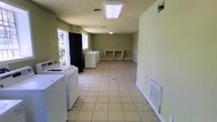 This single-floor laundry facility is designed with practicality and functionality at its core. The space is finished with light beige ceramic tile flooring throughout, providing a clean and durable surface suited to heavy daily use. Walls are painted in a muted sage green, contributing to a calm, utilitarian atmosphere.

Multiple top-load washing machines and dryers are arranged along one side of the room, offering ample capacity for communal laundry needs. A built-in countertop workspace runs along the far wall, providing a convenient surface for folding and sorting laundry. The layout allows for comfortable movement throughout the space.

Overhead fluorescent lighting ensures the room is well-lit, while a window on one end admits natural light and ventilation. A wall-mounted HVAC vent helps regulate the climate within the space. The ceiling features a standard flat finish with a central air return vent.

Overall, the facility is straightforward in design, prioritizing accessibility and ease of use. The open floor plan and organized equipment placement make it a practical and efficient shared laundry space.