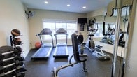 This single-floor fitness room is designed to support a well-rounded workout routine. The space is outfitted with multiple treadmills positioned in a row facing large windows, allowing natural light to fill the room and providing a pleasant view during cardio sessions. An elliptical trainer is situated along the mirrored wall on one side, which effectively amplifies the sense of space and allows users to monitor their form during exercise. A weight bench with attachments occupies the central area, catering to strength training needs. A stability ball adds a versatile option for core and flexibility work. Overhead recessed lighting supplements the natural light from the windows, ensuring the room remains well-lit at all times. A wall-mounted television is positioned for easy viewing from the cardio machines, enhancing the workout experience. The flooring consists of durable rubber matting, appropriate for high-traffic fitness use and providing cushioning underfoot. A wall-mounted fan contributes to ventilation and comfort. The neutral-toned walls and clean layout give the space a practical, no-frills atmosphere focused on functionality.