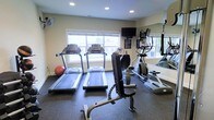 This single-floor fitness room is designed to support a well-rounded workout routine. The space is outfitted with multiple treadmills positioned in a row facing large windows, allowing natural light to fill the room and providing a pleasant view during cardio sessions. An elliptical trainer is situated along the mirrored wall on one side, which effectively amplifies the sense of space and allows users to monitor their form during exercise. A weight bench with attachments occupies the central area, catering to strength training needs. A stability ball adds a versatile option for core and flexibility work. Overhead recessed lighting supplements the natural light from the windows, ensuring the room remains well-lit at all times. A wall-mounted television is positioned for easy viewing from the cardio machines, enhancing the workout experience. The flooring consists of durable rubber matting, appropriate for high-traffic fitness use and providing cushioning underfoot. A wall-mounted fan contributes to ventilation and comfort. The neutral-toned walls and clean layout give the space a practical, no-frills atmosphere focused on functionality.