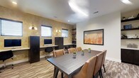This single-floor business center is thoughtfully designed to accommodate a variety of professional needs in a polished, comfortable environment. The space blends functionality with refined aesthetics, creating an atmosphere conducive to both productivity and collaboration.

Along one wall, individual workstations are built into a sleek, dark-finished desk surface, each equipped with a desktop computer and accompanied by ergonomic task chairs. Warm wall sconces illuminate the textured, gold-toned accent wall behind the workstations, adding a sophisticated visual layer to the space. Open shelving above provides convenient storage for books and office supplies, while additional cabinet units offer further organization.

At the center of the room, a substantial dark wood conference table is surrounded by cushioned leather chairs with nailhead trim detail, making it ideal for meetings or collaborative work sessions. Small potted greenery on the table adds a fresh, welcoming touch.

Natural light filters in through horizontal-slatted blinds on the windows, complementing the recessed ceiling lighting. A large framed piece of artwork featuring warm amber and orange tones anchors the opposite wall, enhancing the overall warmth of the interior. Wood-look flooring grounds the space with a clean, contemporary finish, completing this well-balanced and professional environment.
