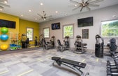 This fitness center offers a comprehensive workout environment spread across a single open floor. The space is thoughtfully designed with a neutral color palette accented by bold yellow-green walls that add energy to the room. Large windows line multiple walls, flooding the interior with abundant natural light and providing pleasant views of surrounding greenery, creating an inviting atmosphere for exercise.

The cardio zone is well-appointed with multiple treadmills and an elliptical machine, each equipped with individual display screens for entertainment during workouts. The strength-training area features a variety of cable and resistance machines designed to target different muscle groups, complemented by a dedicated functional fitness section stocked with stability balls, resistance bands, and other accessories stored on a organized rack.

Flooring throughout the main workout area consists of durable patterned carpet tiles with yellow geometric accents, providing both cushioning and visual interest. Recessed lighting and ceiling fans ensure comfort during intense sessions, while wall-mounted televisions are positioned throughout for convenience. An adjacent corridor provides access to restrooms and features dual water fountains and tasteful artwork, rounding out the facility's amenities with a polished, well-maintained feel.