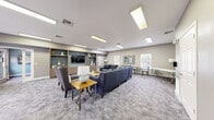 Located at 630 S Peck Dr in Longmont, Colorado, this amenity facility combines a Club Room, Business Center, and Fitness Center into a cohesive, thoughtfully designed space. The Club Room serves as the social centerpiece, featuring a spacious lounge area appointed with a large navy sectional sofa, accent chairs, and a wooden coffee table set atop patterned carpeting. A built-in media wall with warm wood shelving and a flat-screen TV anchors the room, while recessed panel lighting and natural light from surrounding windows create a bright, welcoming atmosphere. Neutral gray walls and contemporary décor give the space a polished, modern aesthetic. Adjacent to the lounge, the Business Center provides a dedicated workspace equipped with a long built-in desk, computer workstation, printer, scanner, and additional office equipment — ideal for productivity needs. The walls here carry a soft blue tone, complementing the overall color palette of the facility. The Fitness Center is well-equipped with treadmills, an elliptical, a stationary bike, a multi-function cable machine, a power rack, a weight bench, and a full dumbbell rack. Wood-look flooring, floor-to-ceiling mirrors, recessed lighting, and a ceiling fan make the exercise area both functional and comfortable for a complete workout experience.
