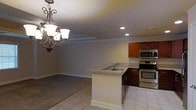 This two-floor space showcases an open-concept layout that seamlessly connects the kitchen and living areas. The kitchen is well-appointed with rich cherry wood cabinetry, stainless steel appliances including a built-in microwave and oven range, and a peninsula island featuring a granite countertop with an integrated sink. The island serves as a functional divider between the kitchen and the adjacent living area, providing both additional prep space and a natural transition point between the two zones.

The flooring transitions from light ceramic tile in the kitchen to carpeting in the living area, creating a subtle distinction between functional and relaxed spaces. Crown molding runs along the ceiling perimeter, adding a refined architectural detail throughout. Recessed lighting in the kitchen provides practical illumination, while a decorative wrought-iron chandelier with frosted glass shades anchors the dining area, adding a touch of elegance. The neutral wall tones and white trim contribute to a clean, cohesive aesthetic throughout the main level, creating a welcoming and functional environment.