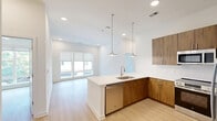 Located at 1351 East Woodlawn Road in Charlotte, NC, this thoughtfully designed 2-bedroom, 2-bathroom residence offers a clean, modern aesthetic across a single floor. The open-concept living area seamlessly connects the dining and kitchen spaces, creating an airy and functional environment bathed in natural light from large windows. The kitchen is a standout feature, showcasing warm wood-tone cabinetry, quartz countertops, a spacious peninsula with an undermount sink, stylish pendant lighting, and a full suite of stainless steel appliances including a refrigerator with ice maker, range, built-in microwave, and dishwasher. Light wood-style flooring flows consistently throughout the entire residence, enhancing the bright and cohesive atmosphere. The primary bedroom is generously sized, offering a ceiling fan and ample natural light, and connects to a well-appointed en-suite bathroom featuring a single vanity with quartz countertop and a glass-enclosed walk-in shower. The second bedroom is equally bright, with a large window overlooking the neighborhood. The second bathroom includes a tiled tub-shower combo and a wood-tone vanity. Additional conveniences include multiple closets with built-in shelving, a dedicated laundry area, and a utility room.