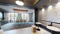 This single-floor space is thoughtfully designed to balance professional functionality with contemporary elegance. It is divided into two distinct yet visually connected zones: a glass-enclosed conference room and an open lounge area, each finished to a high standard.

The conference room is anchored by a long white table surrounded by ergonomic mesh chairs and illuminated by a striking globe chandelier with brass accents. A coffered ceiling detail frames the fixture, adding architectural depth. Floor-to-ceiling windows on two sides flood the room with natural light and provide views of lush outdoor greenery, creating an inviting and productive atmosphere.

Separating the two zones is a dramatic accent wall clad in chevron-patterned stone tile in neutral tones, serving as a focal point that bridges the spaces. The lounge area features built-in upholstered banquette seating with dark wood bases, complemented by round bistro tables and cylindrical ottomans finished in white leather and natural wood veneer. Large-format light tile flooring extends throughout this zone, contrasting with the textured charcoal wall panels. Full-length mirror panels with grid framing visually expand the lounge, reflecting the pendant lighting above and enhancing the overall sense of depth and sophistication.