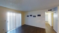 This single-floor space presents a clean, straightforward layout with a welcoming main living area as its centerpiece. The room features dark hardwood flooring that contrasts cleanly against the white baseboards and light-colored walls, creating a grounded, polished aesthetic. A textured popcorn ceiling adds a classic residential character to the space.

Natural light is a notable feature of this room, introduced through a large floor-to-ceiling window dressed with vertical blinds, allowing for adjustable light control throughout the day. The blinds, when open, suggest access to an outdoor area or patio.

The walls are kept minimal and neutral, currently adorned with a series of four framed artworks arranged in a horizontal line, demonstrating the wall space available for personalization. The room opens into an adjacent area, hinting at a connected layout that promotes a sense of flow between spaces.

Overall, the space offers a functional and adaptable environment. Its combination of dark flooring, ample natural light, and neutral tones provides a versatile foundation suitable for a variety of interior design approaches.