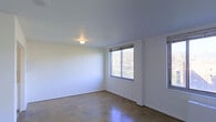 This single-floor space presents a straightforward and functional layout defined by clean lines and a neutral aesthetic. The interior is finished with smooth white walls and flat ceilings throughout, creating a bright and open atmosphere. Underfoot, distinctive patterned tile flooring adds a subtle visual texture to the space, providing a durable and easy-to-maintain surface across the main living area.

Natural light plays a key role in the ambiance of this space. Windows fitted with horizontal blinds allow for adjustable light control while offering views of the outdoors, where mature trees and surrounding greenery are visible — lending a calm, natural backdrop to the interior. A flush-mount ceiling light fixture provides overhead illumination, ensuring the space remains well-lit regardless of the time of day.

The open, unobstructed layout of the main room offers flexibility for a variety of uses and configurations. The walls are clean and unmarked, providing a blank canvas for personalization. The overall design is minimal and utilitarian, emphasizing practicality and ease of use. This space is well-suited for those seeking a simple, move-in-ready environment with a modest and unpretentious character.